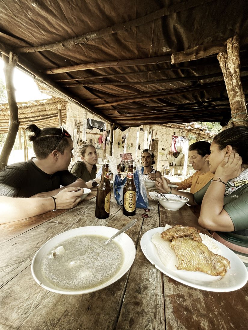 Riders sharing lunch at a long wooden table under a thatched roof